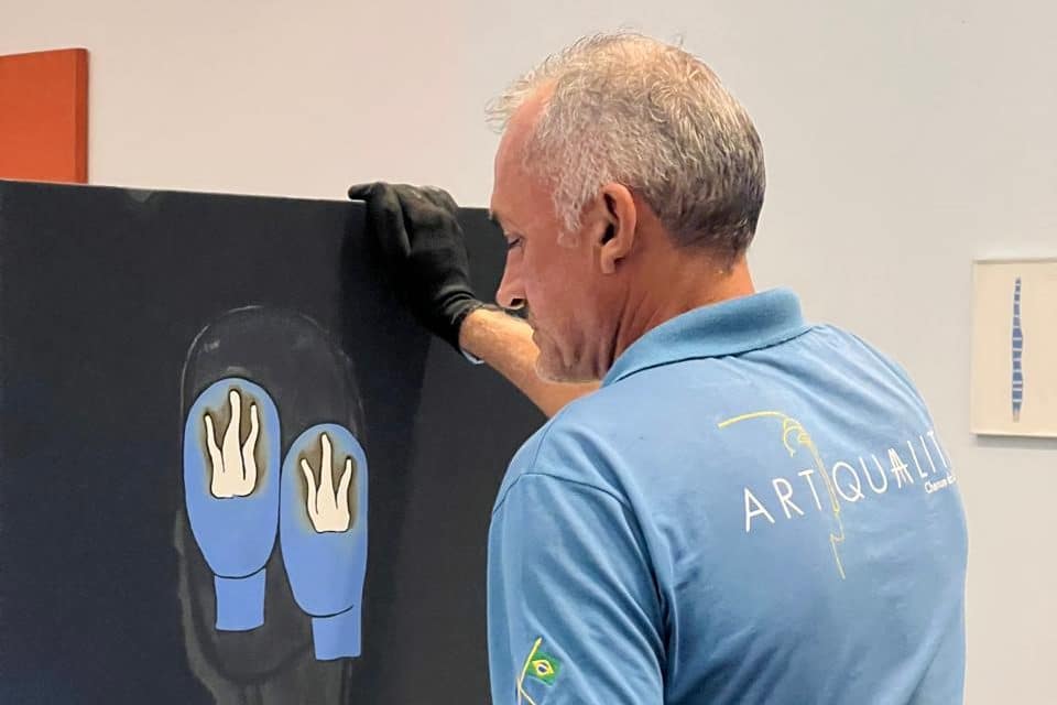 Técnico de luva preta apoia uma tela escura com duas formas azuis durante a montagem da exposição