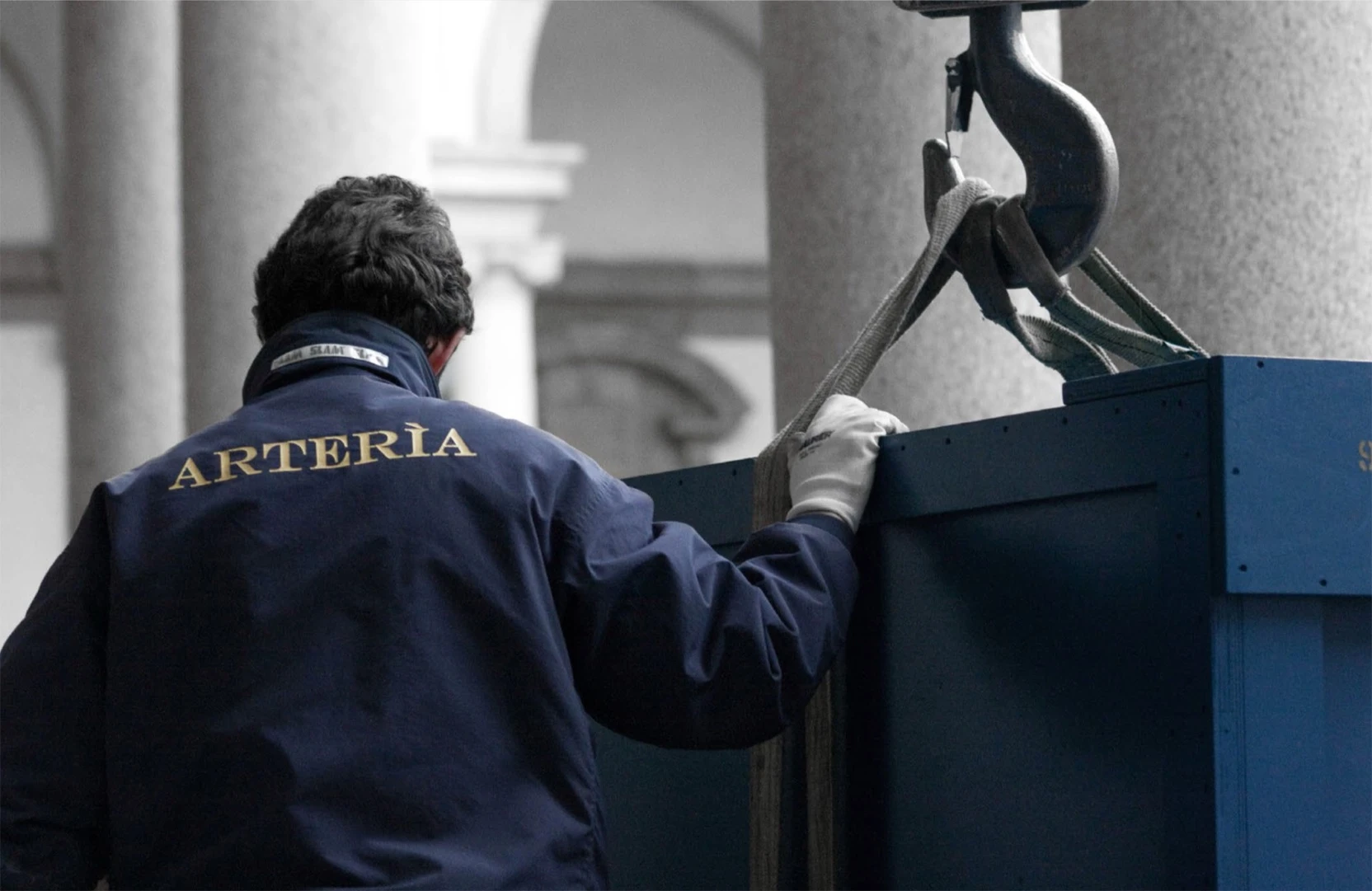 Técnico de jaqueta Arterìa apoia a mão numa caixa azul de transporte de obras de arte, suspensa por um gancho de guindaste no pátio de um palácio