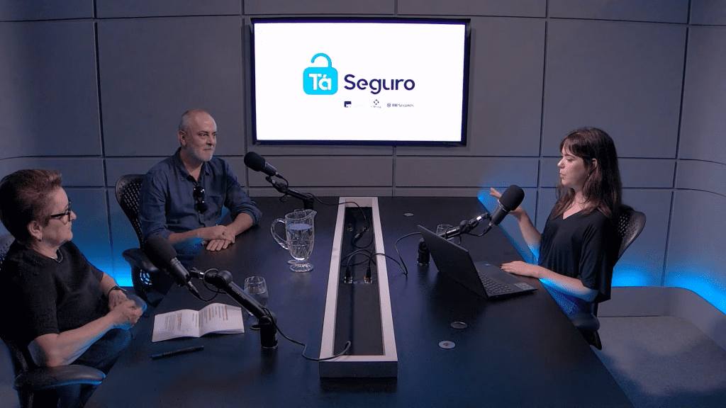 Três participantes conversam ao redor da mesa de um estúdio de podcast, diante de microfones e de uma tela com o logo do programa