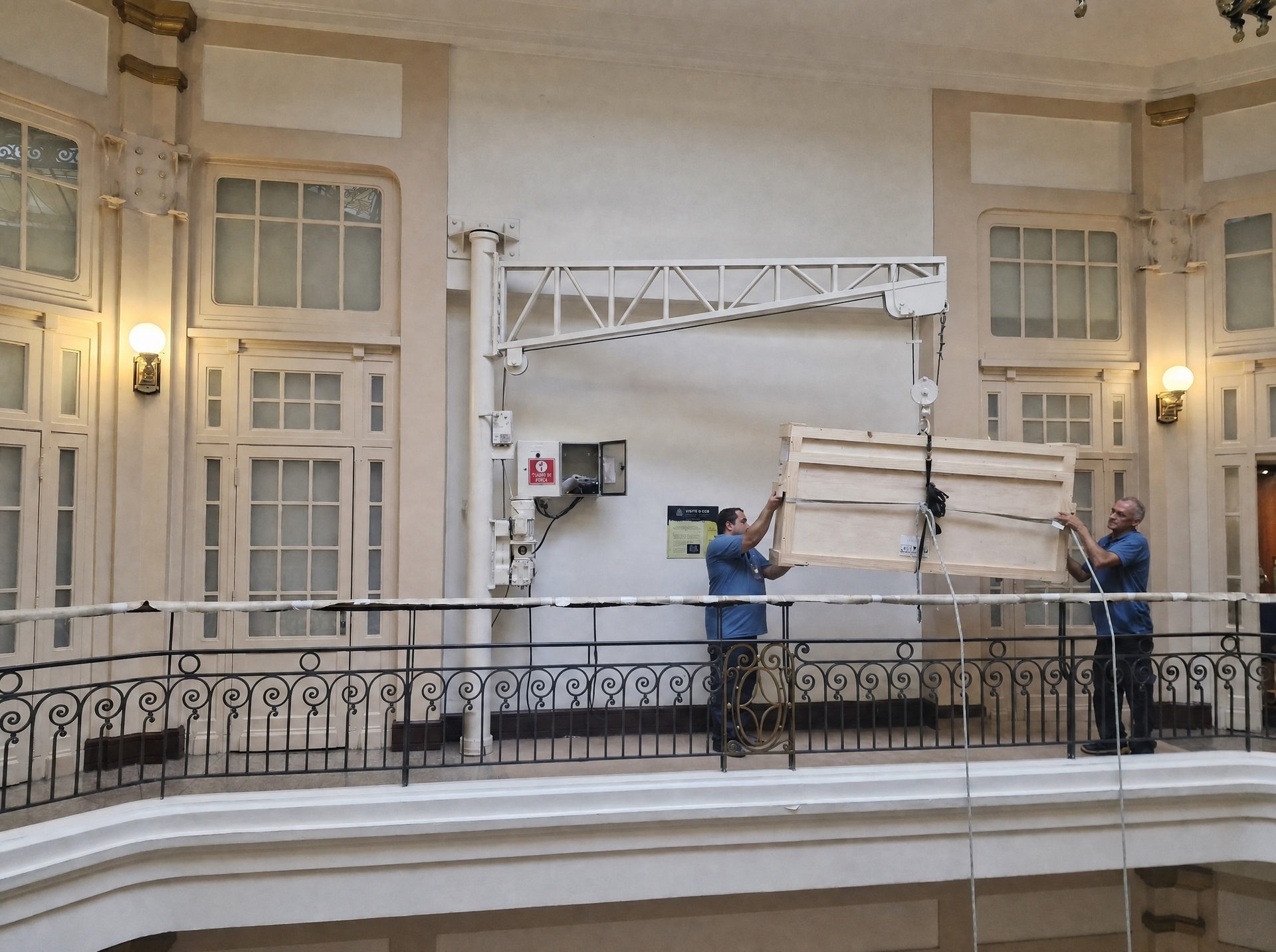 Dois operadores prendem a cinta de içamento a uma caixa embalada e cintada, sob o pórtico móvel, na varanda de um edifício de frente para o mar