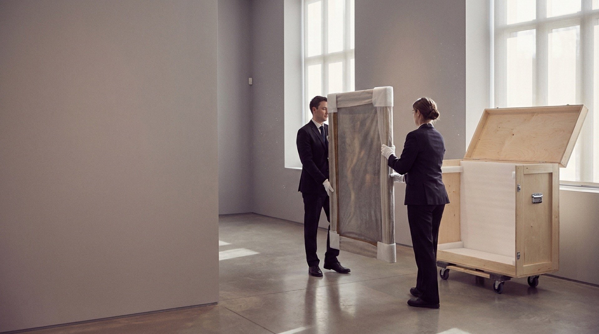 Two white-gloved handlers carry a protected painting to an open wooden crate in a museum room